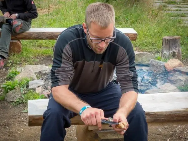 Mann schnitzt mit einem Messer an einem Holzstück, Feuerstelle im Hintergrund