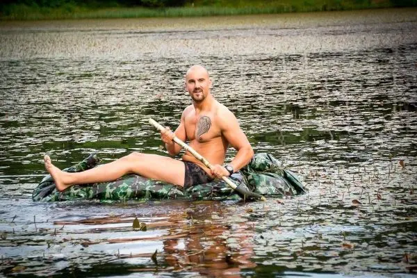 Mann sitzt auf einer camouflagefarbenen Schwimmhilfe im Wasser und hält einen Stock