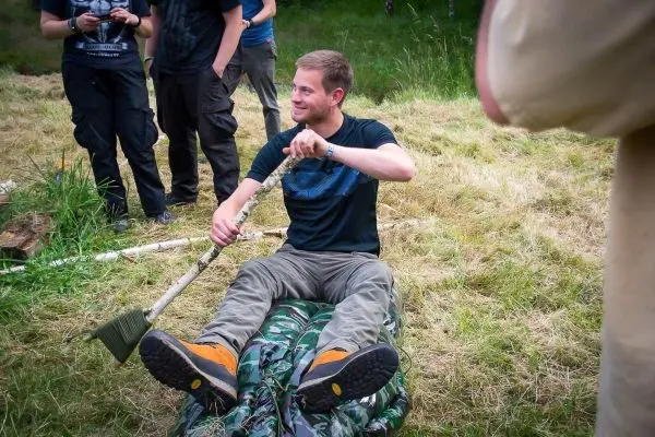 Mann sitzt auf einer camouflagefarbenen Unterlage und hält einen Stock in der Hand