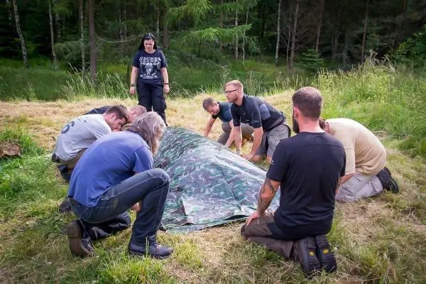 Sechs Personen bauen gemeinsam eine Tarp-Notunterkunft aus einer camouflierte Plane auf