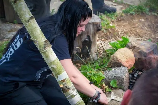 Zunder wird mit einem Messer vorbereitet, während ein Feuerplatz im Hintergrund sichtbar ist