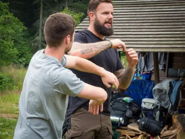 Zwei Personen demonstrieren eine Technik im Freien, Rucksäcke und Ausrüstung im Hintergrund