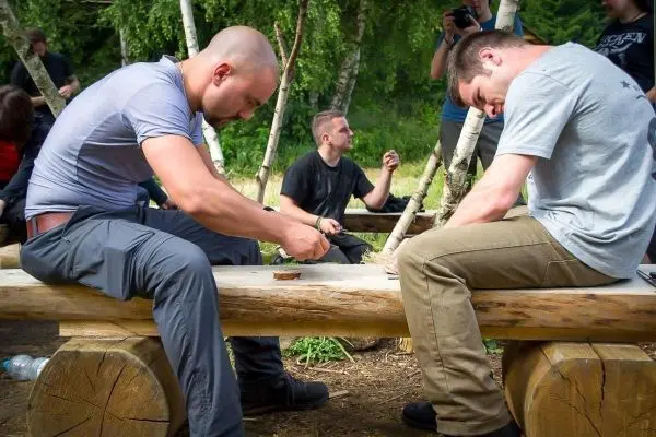 Zwei Personen schnitzen Holz auf einer Baumstamm-Bank im Freien