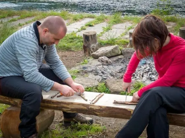 Zwei Personen schnitzen mit Messern an einem Holzstück auf einer Bank