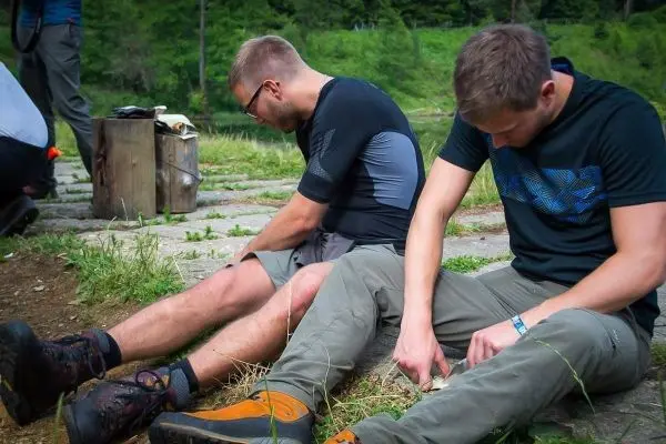 Zwei Personen sitzen am Boden und schnitzen mit Messern an Holzstücken