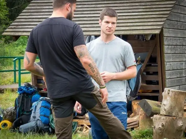 Zwei Personen stehen vor einer Holzhütte, Rucksäcke liegen im Hintergrund