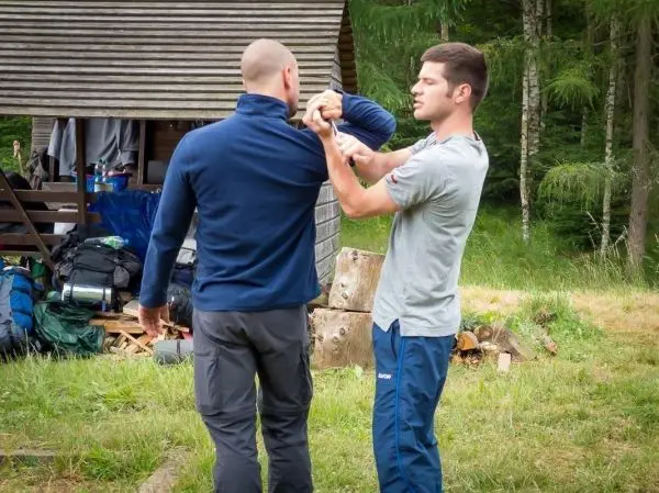 Zwei Personen üben eine Selbstverteidigungstechnik im Freien nahe einer Hütte