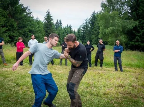 Zwei Personen üben einen Nahkampfgriff, Zuschauer beobachten im Hintergrund