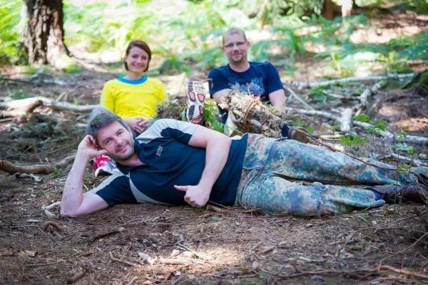 Drei Personen sitzen und liegen im Wald auf dem Boden zwischen Ästen