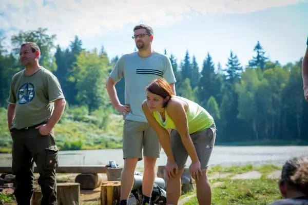 Drei Personen stehen am Ufer eines Gewässers, eine Person beugt sich nach vorne