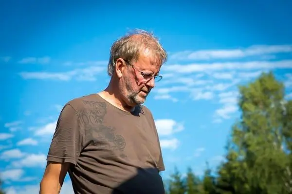 Ein Mann steht nachdenklich im Freien, umgeben von Bäumen und blauem Himmel