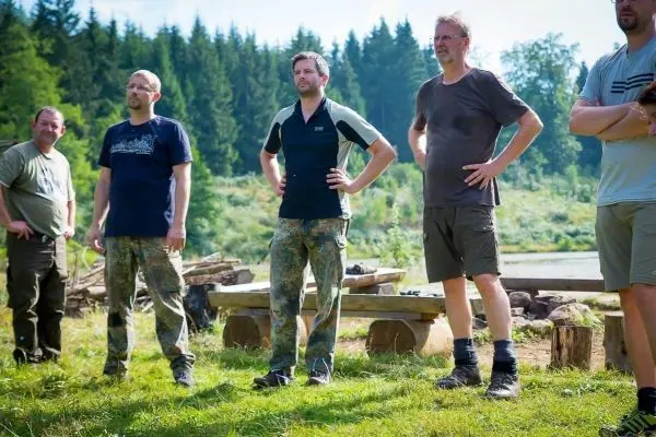 Fünf Personen stehen in einer Wiese neben einem Holzstapel und einem Gewässer