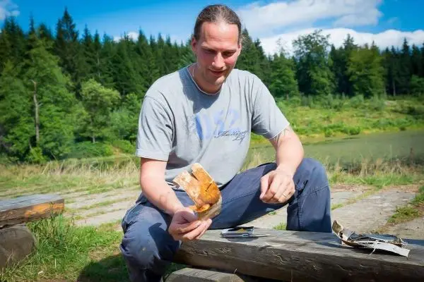 Mann hält ein Stück getrockneten Fisch in der Hand, sitzt an einem Holzstamm