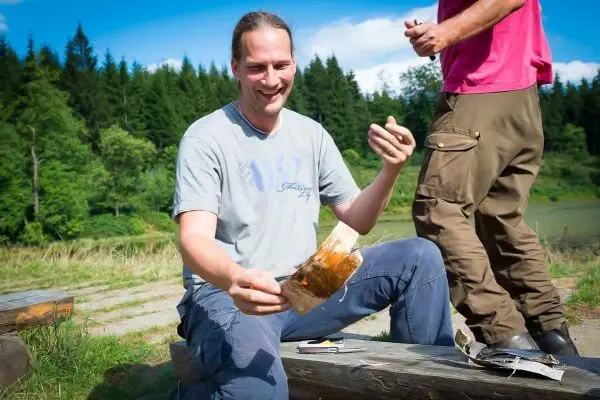 Mann hält ein Stück Holz in der Hand, sitzt auf einer Bank im Freien