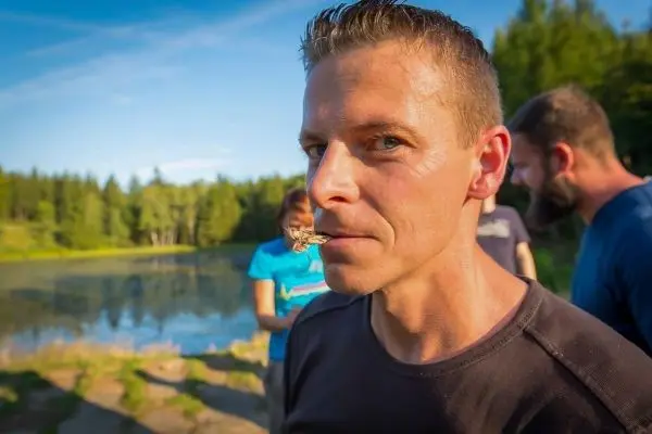 Mann hält einen Insekt zwischen den Lippen, im Hintergrund ein Gewässer und Bäume