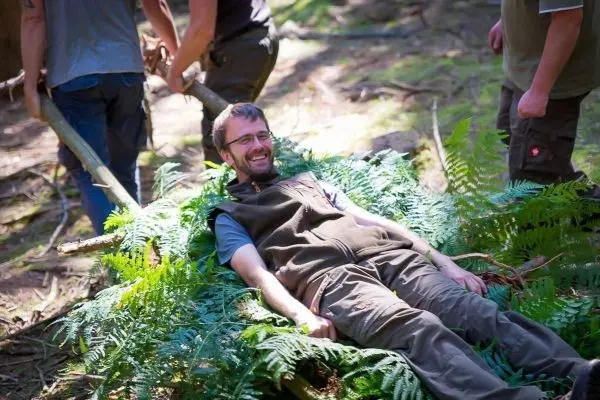 Mann liegt auf einem Bett aus Farnen, während andere Personen Holzstücke halten