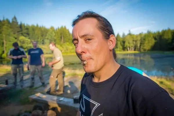 Mann mit einem Stock im Mund steht am Ufer eines Gewässers, im Hintergrund sind weitere Personen