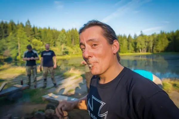 Mann mit einem Stück Holz im Mund steht am Ufer eines Gewässers