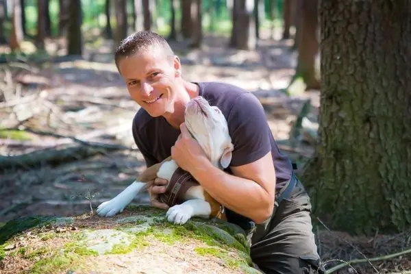Mann sitzt auf einem Baumstamm und umarmt einen Hund im Wald