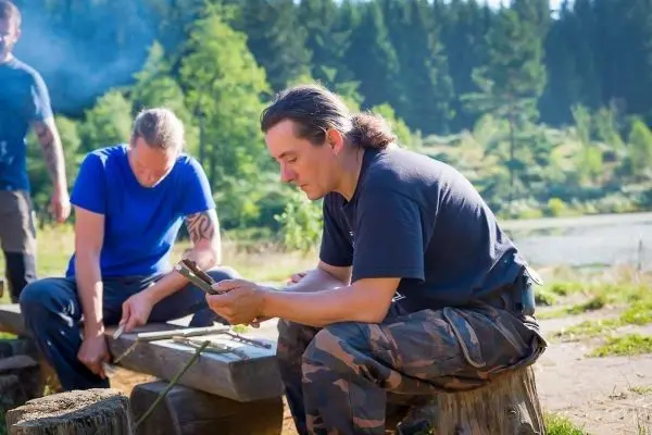 Mann sitzt auf einem Baumstumpf und betrachtet ein Messer, während ein anderer an einem Tisch arbeitet