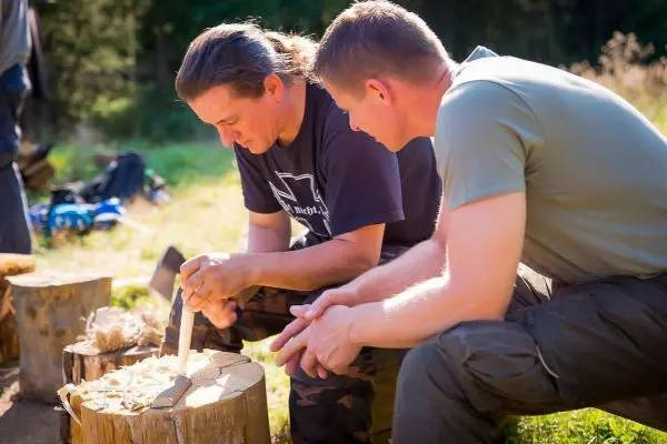 Messer bearbeitet Holzstück auf einem Baumstumpf, zwei Personen beobachten aufmerksam
