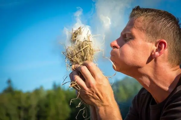 Person bläst in ein Bündel aus trockenem Gras und erzeugt Rauch beim Feuer machen