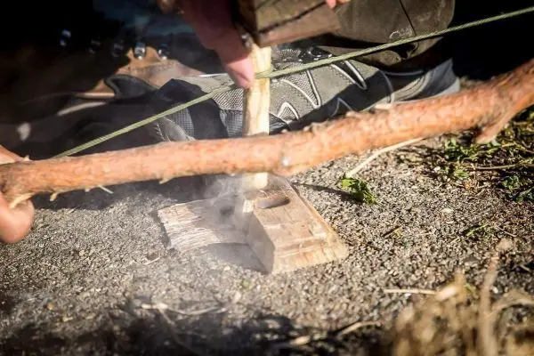 Person erzeugt Rauch mit einem Holzstück auf einer Unterlage beim Survival Training