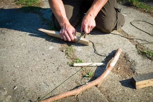 Person schnitzt mit einem Messer einen Stock auf dem Boden