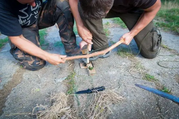 Zwei Personen bauen eine Feuerbohrmaschine mit Holzstücken und Zunder