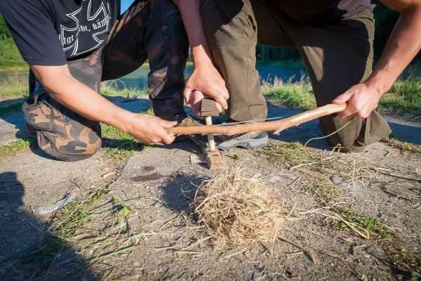 Zwei Personen nutzen einen Feuerbohrer, um Zunder zu entzünden