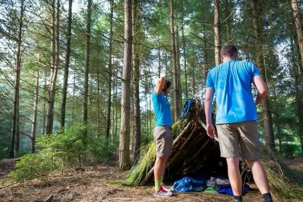 Zwei Personen stehen vor einer selbstgebauten Tarp-Notunterkunft im Wald