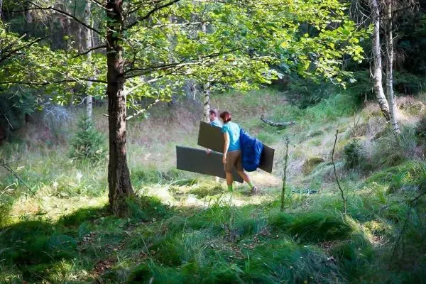 Zwei Personen tragen große Platten durch einen Wald mit hohem Gras