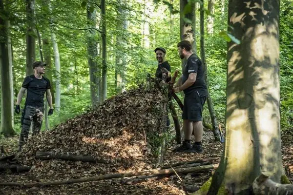 Drei Personen bauen eine Laubhütte aus Ästen und Blättern im Wald