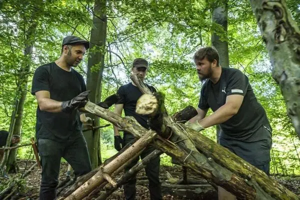 Drei Personen konstruieren eine Holzkonstruktion im Wald mit Ästen und Schnüren
