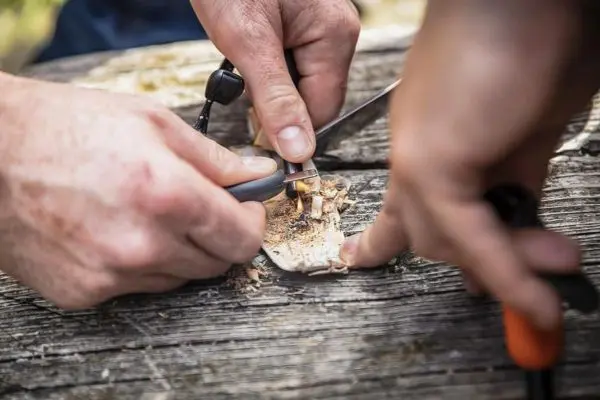 Ein Messer schneidet in ein Stück Holz, während eine Hand Zunder vorbereitet