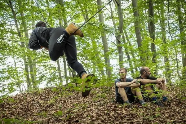 Ein Teilnehmer schwingt an einer Seilbrücke über den Waldboden, während zwei andere sitzen