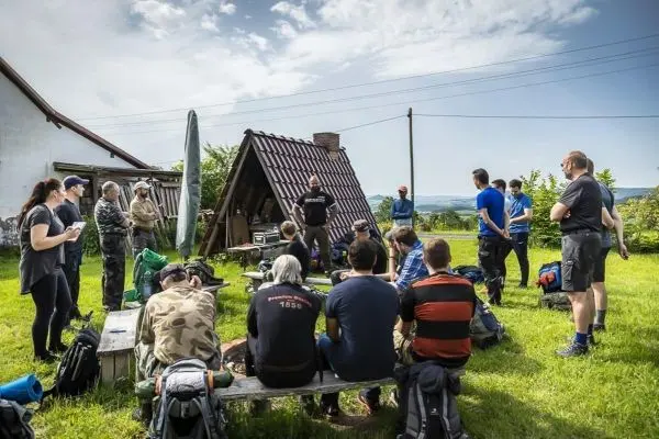 Gruppe hört einem Vortrag unter einem Dachvorsprung zu, Rucksäcke liegen auf Bänken