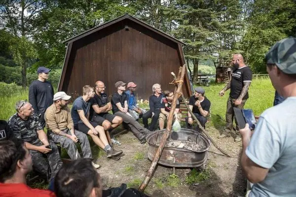 Gruppe sitzt um ein Feuer in einer Feuerschale, Holzunterstand im Hintergrund
