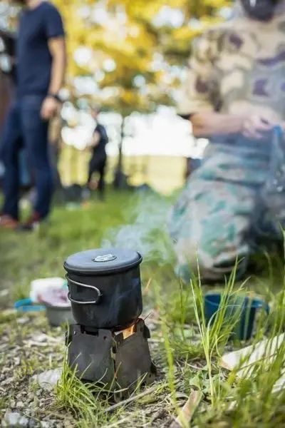Hobo-Kocher mit Kessel auf einem Untergrund aus Gras, Rauch steigt auf