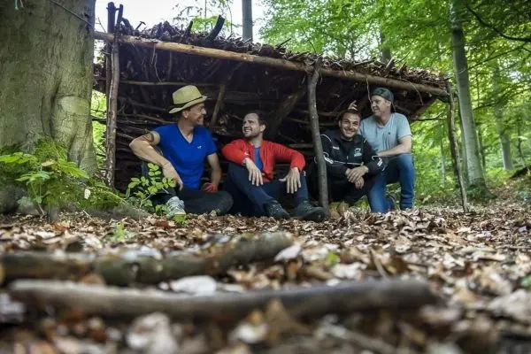 Holzkonstruktion als Notunterkunft im Wald, vier Personen sitzen davor auf dem Boden