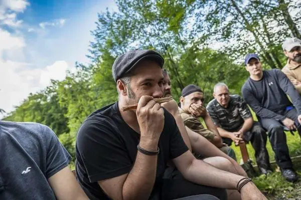 Mann hält Holzstück vor Mund und spricht zu Gruppe im Freien