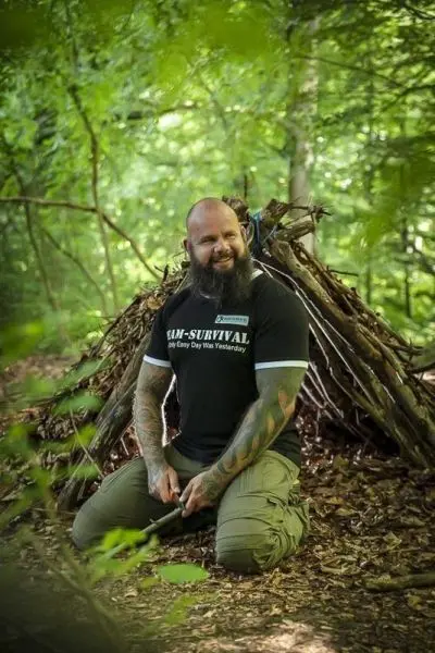 Mann kniet vor einer Laubhütte und hält ein Messer in der Hand