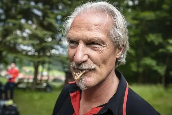 Mann mit grauen Haaren und Schnurrbart hält einen Holzspan im Mund