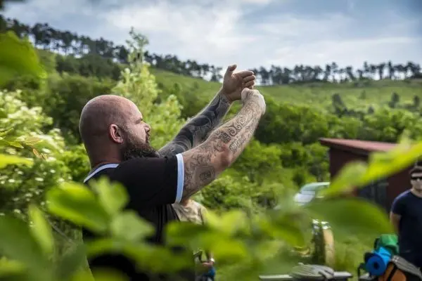 Mann mit tätowierten Armen zeigt eine Geste in einer grünen Umgebung