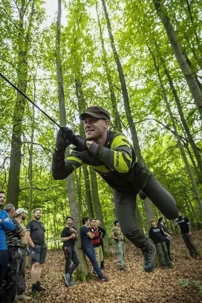 Mann schwingt an einem Seil durch den Wald, Zuschauer beobachten aufmerksam