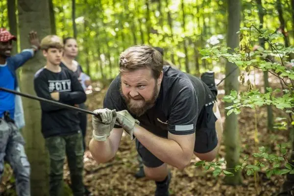 Mann zieht sich an einem Seil durch den Wald, Zuschauer beobachten