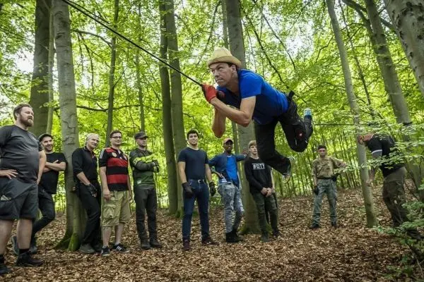 Person schwingt an Seil zwischen Bäumen, Gruppe schaut zu beim Survival Training