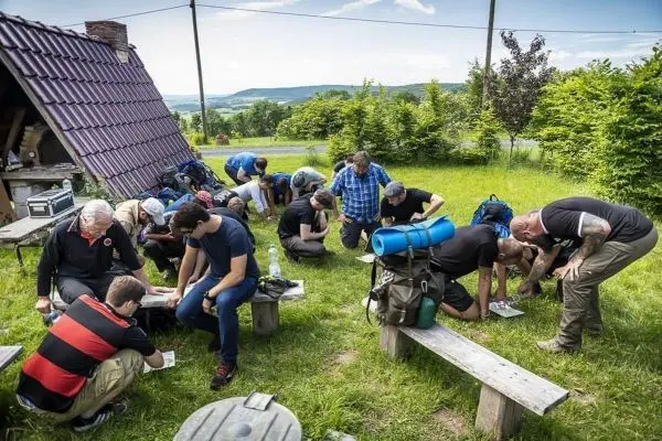 Teilnehmende sitzen an Bänken und schreiben auf Papier, Rucksäcke liegen daneben