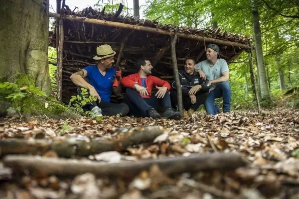 Vier Personen sitzen entspannt unter einer selbstgebauten Holzunterkunft im Wald