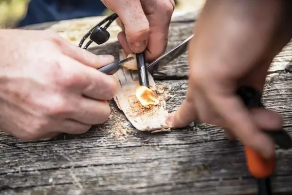 Zwei Hände bearbeiten einen Holzstück mit einem Messer und einem Feuerstahl